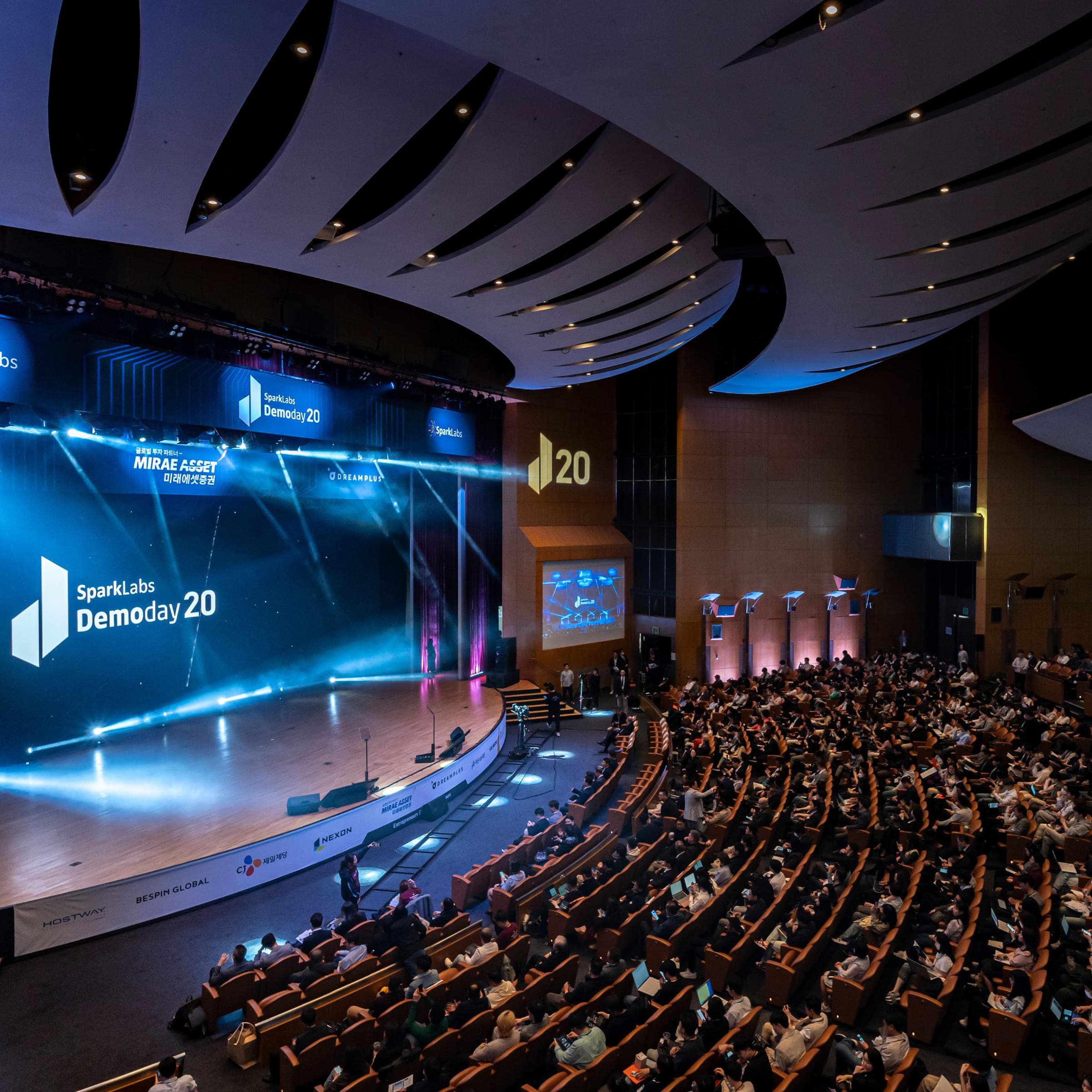 A full auditorium at SparkLabs Demoday 20, the lit stage and its screens facing banked rows of seats