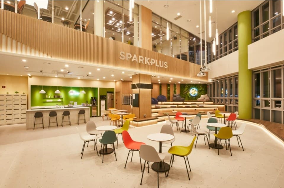 The event hall at SparkPlus Seolleung 3 in Seoul: a double-height space with the SPARKPLUS name on a timber gallery, a coffee bar, tiered seating and café tables with coloured chairs